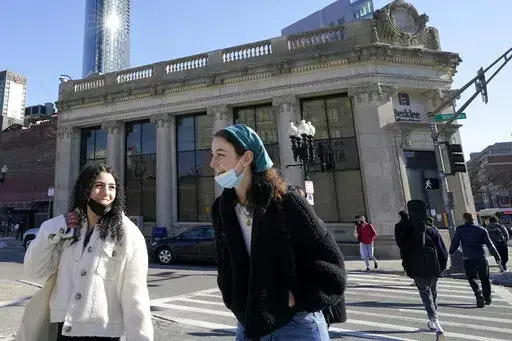 Passers-by wear masks under their chins as they chat with one another while crossing a street, in Boston, Wednesday, Feb. 9, 2022. Students and staff at public schools in Massachusetts will no longer be required to wear face coverings while indoors starting Feb. 28, state officials said Wednesday, Feb. 9, 2022. (AP Photo/Steven Senne, File)