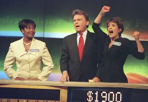 Johnna Goodwin, left, watches as Donna Handel, right, reacts after winning on "Wheel of Fortune," with host Pat Sajak during a taping of the game show in Philadelphia on April 17, 1999. The game show was in Philadelphia for the weekend taping a week's worth of shows. Ryan Seacrest will replace the retiring Sajak as host of “Wheel of Fortune.” (AP Photo/William Thomas Cain, File)