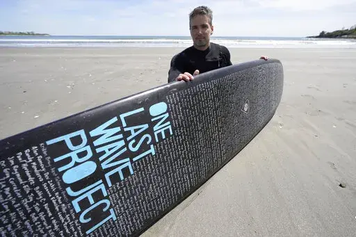 Dan Fischer, of Newport, R.I., sits for a photograph with his surfboard on Easton's Beach, in Newport, Wednesday, May 18, 2022. Fischer, 42, created the One Last Wave Project in January 2022 to use the healing power of the ocean to help families coping with a loss, as it helped him following the death of his father. Fischer places names onto his surfboards, then takes the surfboards out into the ocean as a way to memorialize the lost loved ones in a place that was meaningful to them. (AP Photo