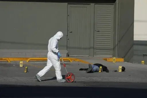An attacker lies dead on his back in the parking lot of a convenience store as a police investigator uses a measuring wheel while collecting evidence from a crime scene where a municipal policeman was shot dead, in Celaya, Mexico, Feb. 28, 2024. The policeman had been driving his wife to work when cartel gunmen opened fire on their car. The policeman killed one attacker before dying. (AP Photo/Fernando Llano, File)