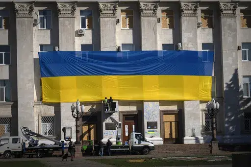 Municipal workers wearing protective vests and helmets decorate the wall of the regional administration building with a huge national flag to mark one year since the Ukrainian troops cleared the city from the Russian army, in Kherson, Ukraine, Friday, Nov. 10, 2023. (AP Photo/Efrem Lukatsky)