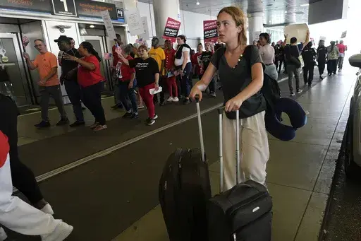 A traveler walks through the departure area as airline catering workers who are employed by Gate Gourmet picket with their supporters, calling for a new union contract with raises and affordable health insurance, Wednesday, July 3, 2024, at Miami International Airport in Miami. A long Fourth of July holiday weekend is expected to create new travel records. (AP Photo/Lynne Sladky)