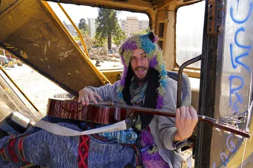 At Atmaa Das, 28, who is homeless from Alabama, strums his guitar while singing parts of Woody Guthrie's anthem, "This Land Is Your Land" while sitting in a piece of construction equipment left behind at People's Park in Berkeley, Calif., on Aug. 16, 2022. The three-acre site's colorful history, forged from University of California, Berkeley's seizure of the land in 1968, has been thrust back into the spotlight by the school's renewed effort to pave over People's Park as part of a $312 million p