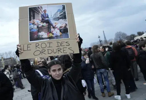A woman holds a placard depicting French President Emmanuel Macron sitting on garbage cans that reads, "king of trash" during a protest in Paris, Friday, March 17, 2023. Protests against French President Emmanuel Macron's decision to force a bill raising the retirement age from 62 to 64 through parliament without a vote disrupted traffic, garbage collection and university campuses in Paris as opponents of the change maintained their resolve to get the government to back down. (AP Photo/Lewis Jol